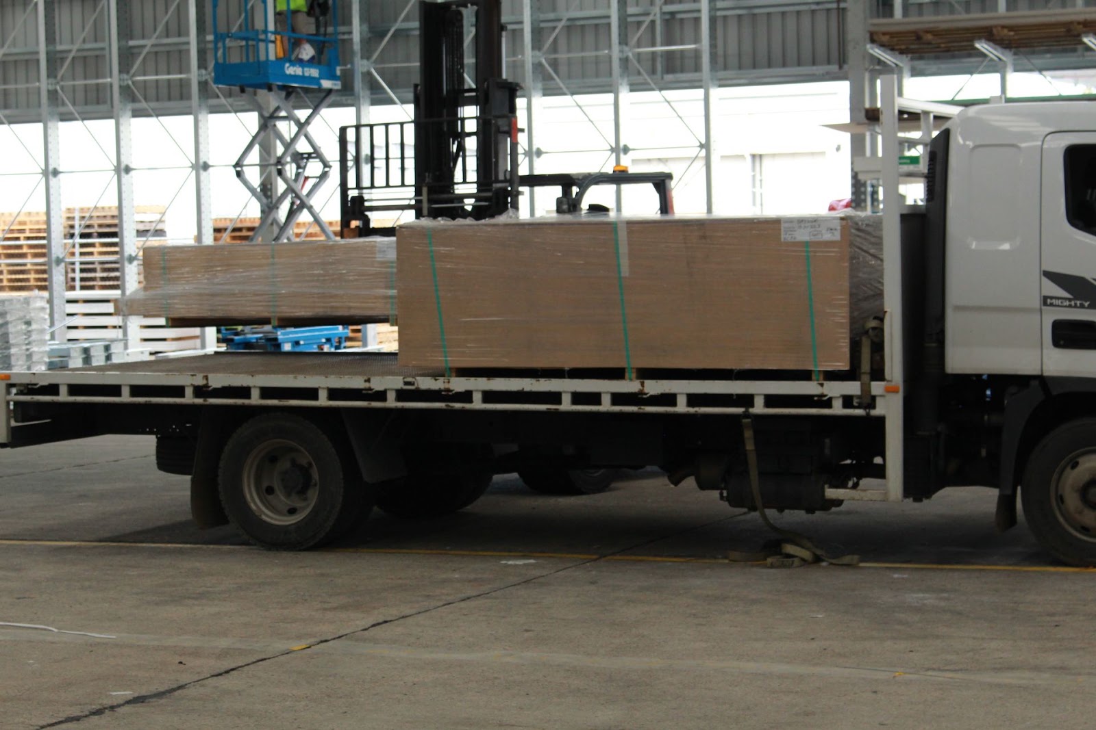 Forklift lowering shrink-wrapped bundles of board onto a Hyundai Mighty flatbed truck, side view inside the warehouse.