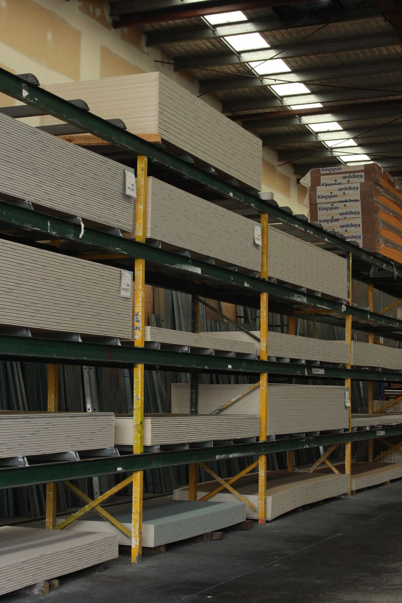 Green and yellow racking stacked with plasterboard sheets, Kingspan insulation boxes on the top bay, skylights above.