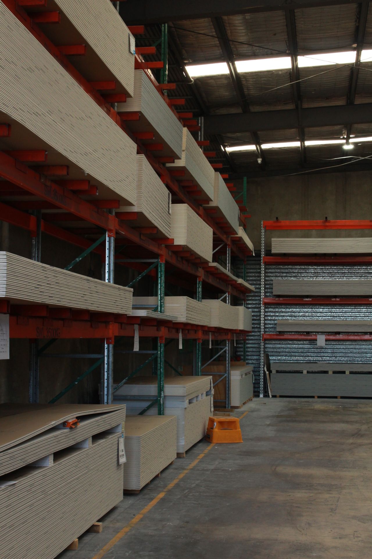 Portrait of tall orange racking loaded with plasterboard, a second rack of sheets at the back wall.