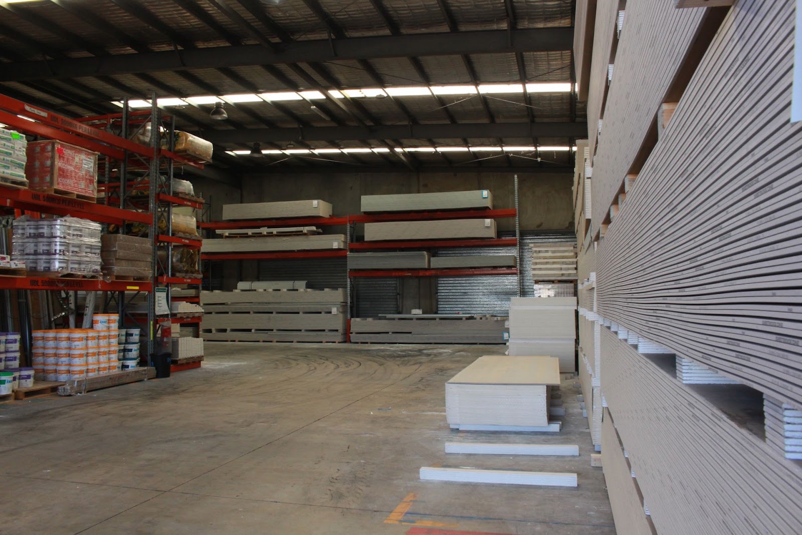 Wide warehouse view: pallet racking of compounds and boxes at left, cantilever racks of sheets at the back, a wall of st